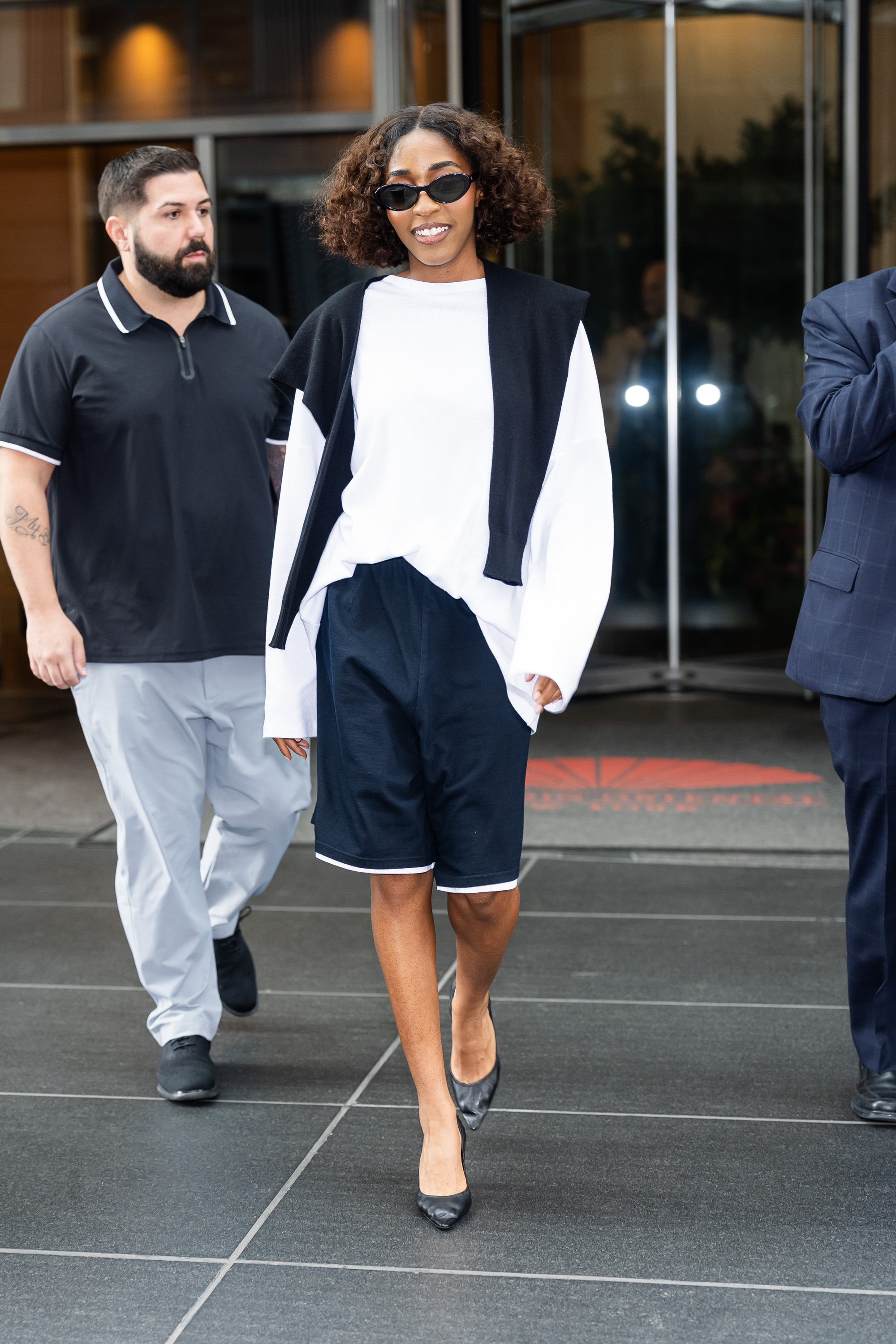 a black woman with brown, shoulder length curly hair photographed walking down the street in a white sweatshirt with navy blue athletic shorts and black heels