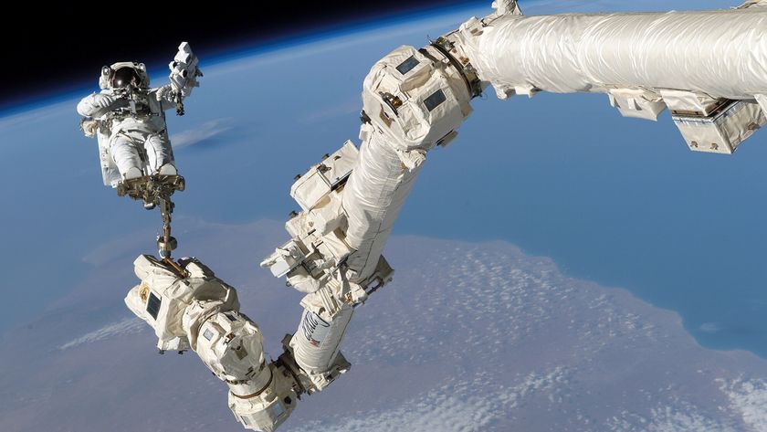 An astronaut in a white suit sits on a large white robotic arm floating above the Earth in space