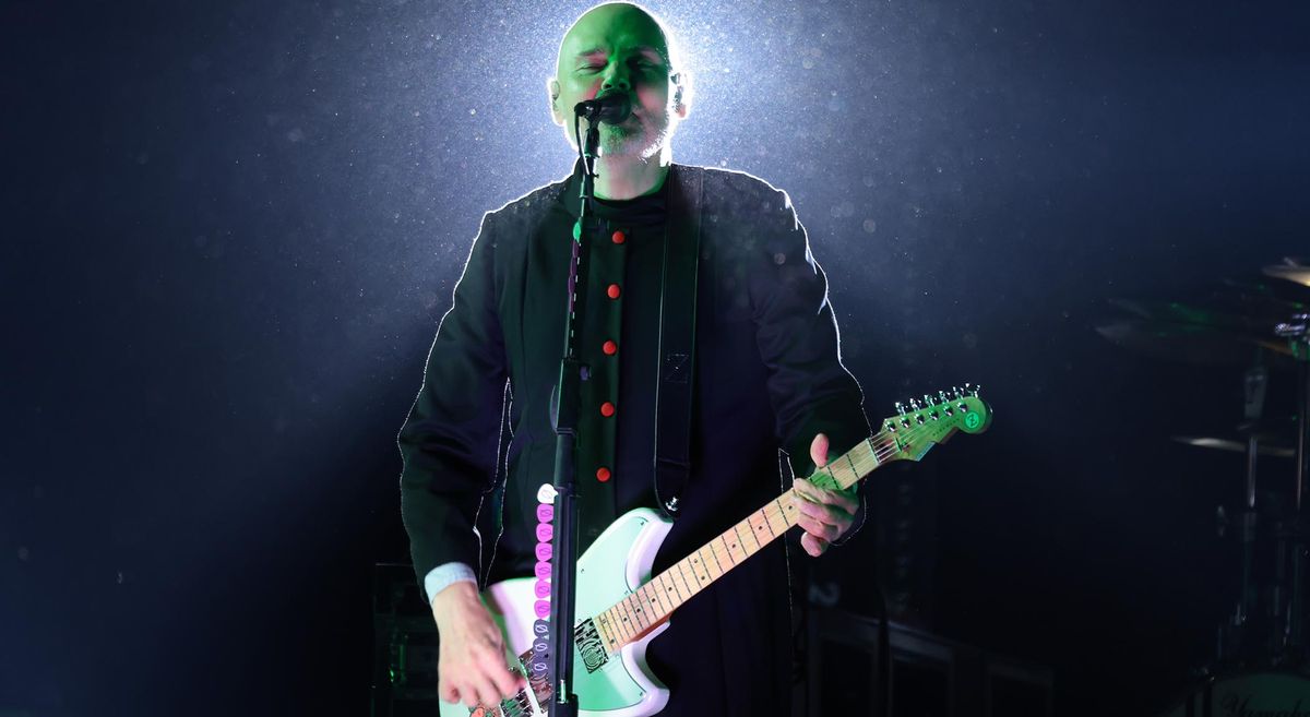 Billy Corgan wears black and plays his Reverend signature guitar onstage with the Smashing Pumpkins, with a stage light illuminating him from behind.