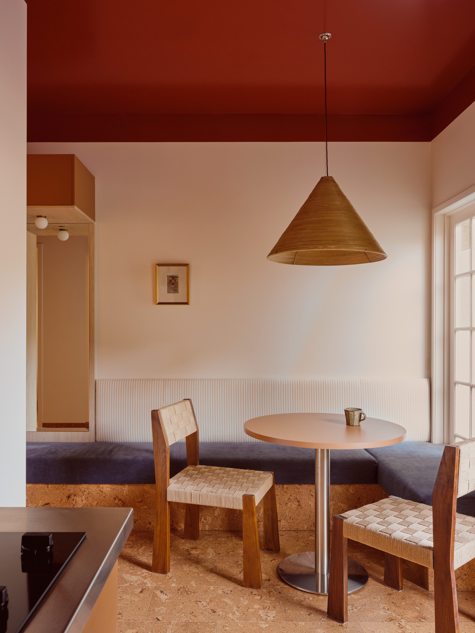 A dining nook with white walls and a red ceiling. There is a built-in bench with blue cushions, a round table, and wooden and white chairs.