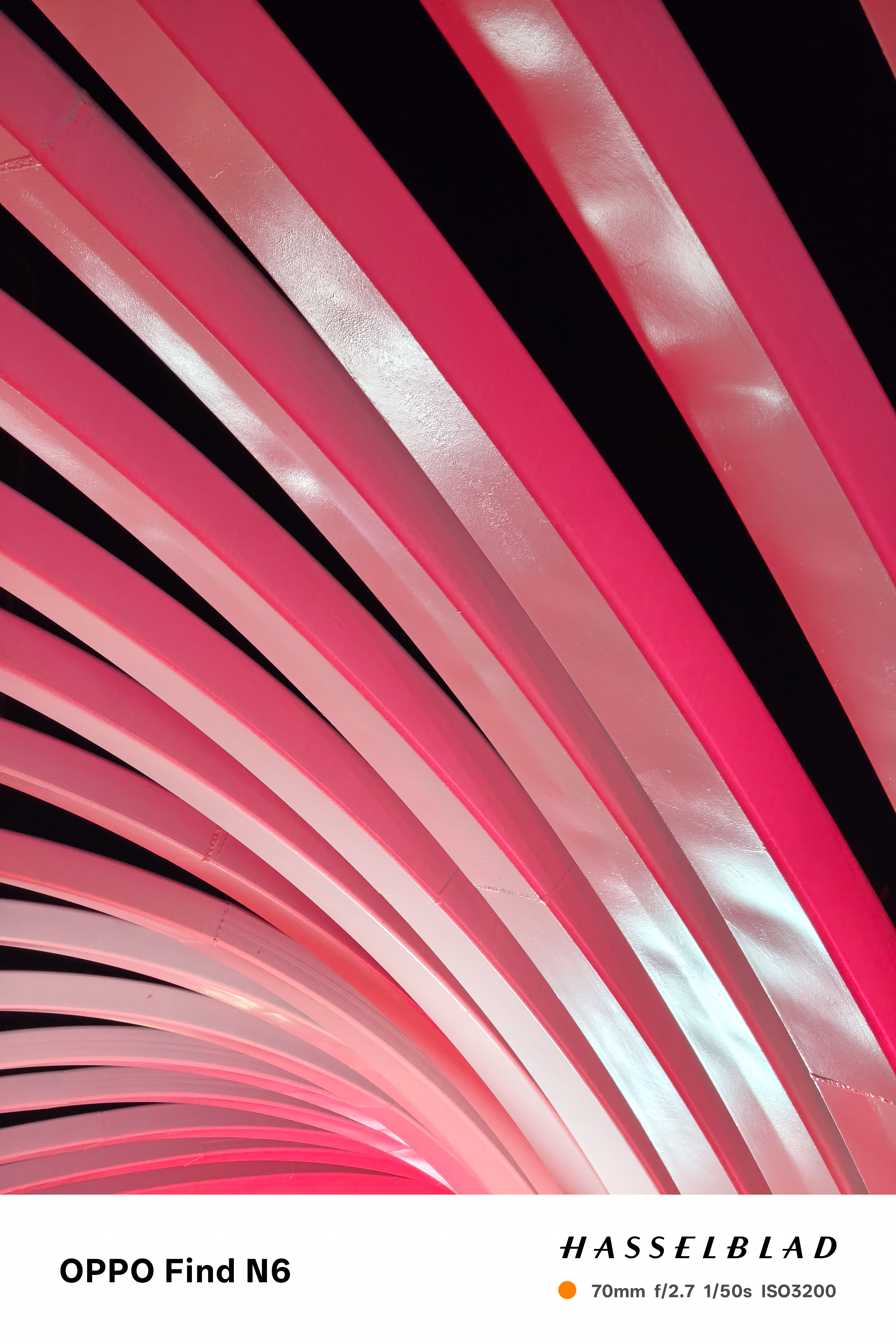 An abstract, close-up shot of a modern sculpture made of multiple curved, pink metallic slats. The slats are lit from the side, creating a gradient of light and shadow that emphasizes their sleek, architectural lines against a black background.