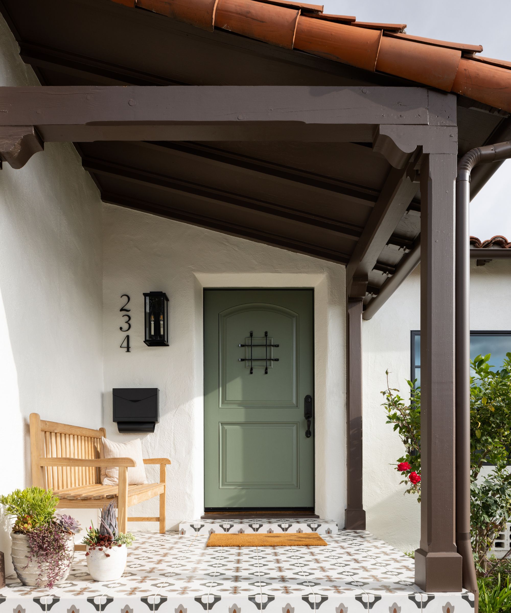Spanish style home with terracotta tiled roof, white and black tiled floor, green door, wood bench and welcome mat