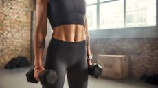 An athletic woman standing and holding two dumbbells with her abs on show