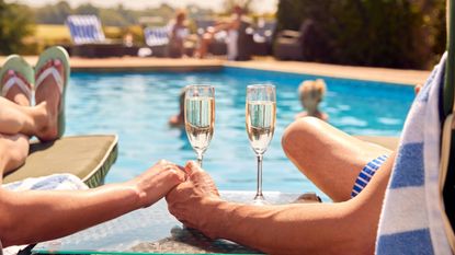 Close up of couple holding hands and champagne glasses with swimming pool in background
