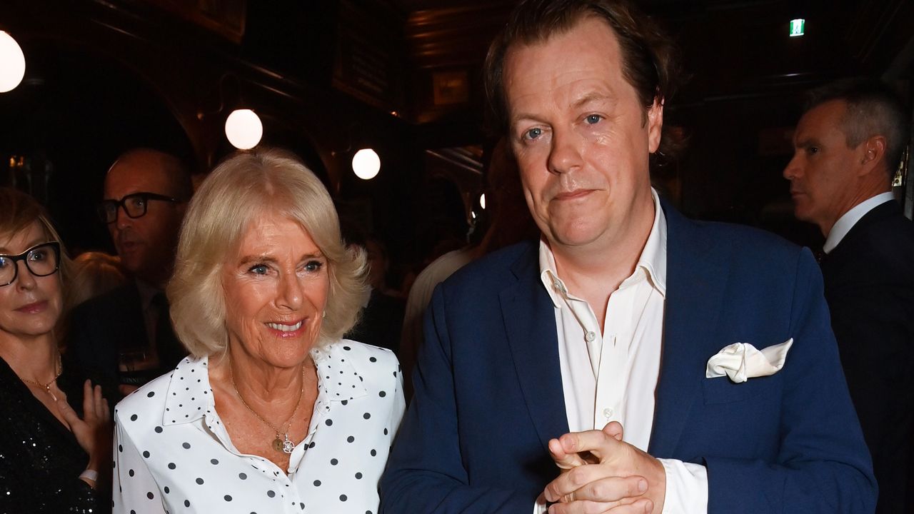 Tom Parker Bowles wearing a blue suit standing next to his mother Queen Camilla who is smiling and wearing a white polkadot dress