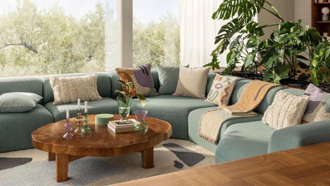 L-shaped blue sofa sectional with white throw pillows; a wooden coffee table with candles and vases; in the corner, a houseplant; all looking onto windows with a garden view. 