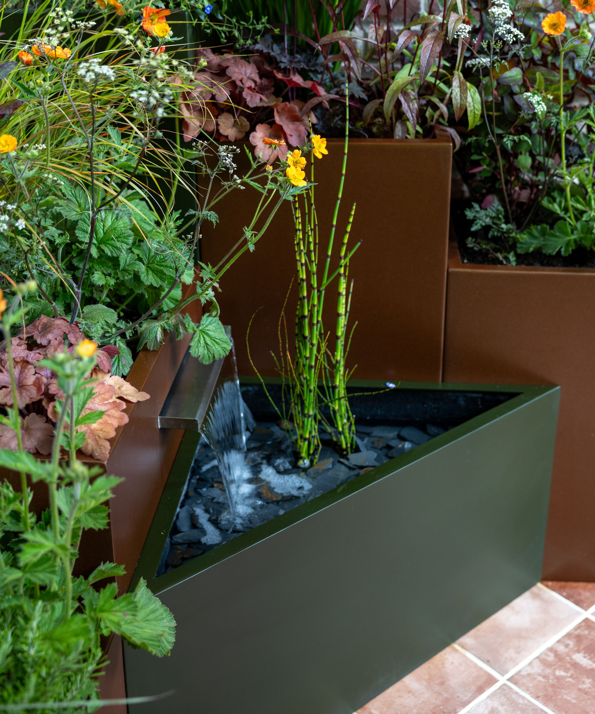 triangular raised bed with plants, positioned in front of more raised beds planted with summer flowers such as geum and heuchera