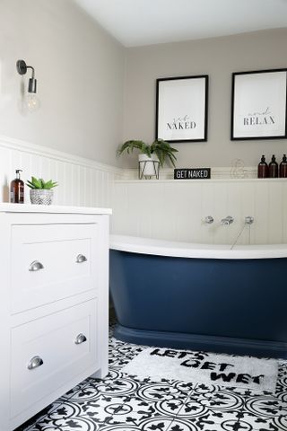 Cream bathroom with panelled walls, monochrome patterned floor tiles, blue roll top bath and Shaker-style grey vanity unit