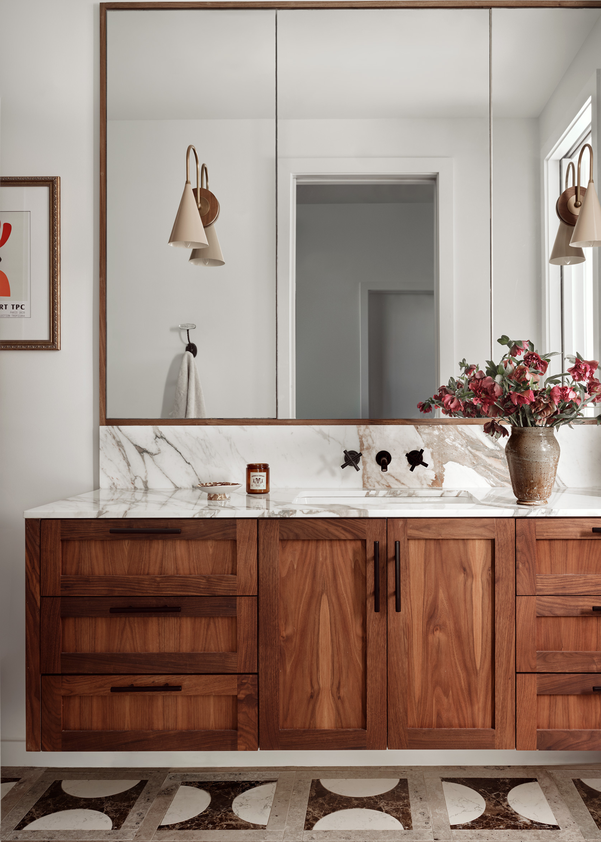 A bathroom with patterned floor tiles a floating cabinet, a marble backsplash, mirrored cabinets, a vase of flowers and a glass candle