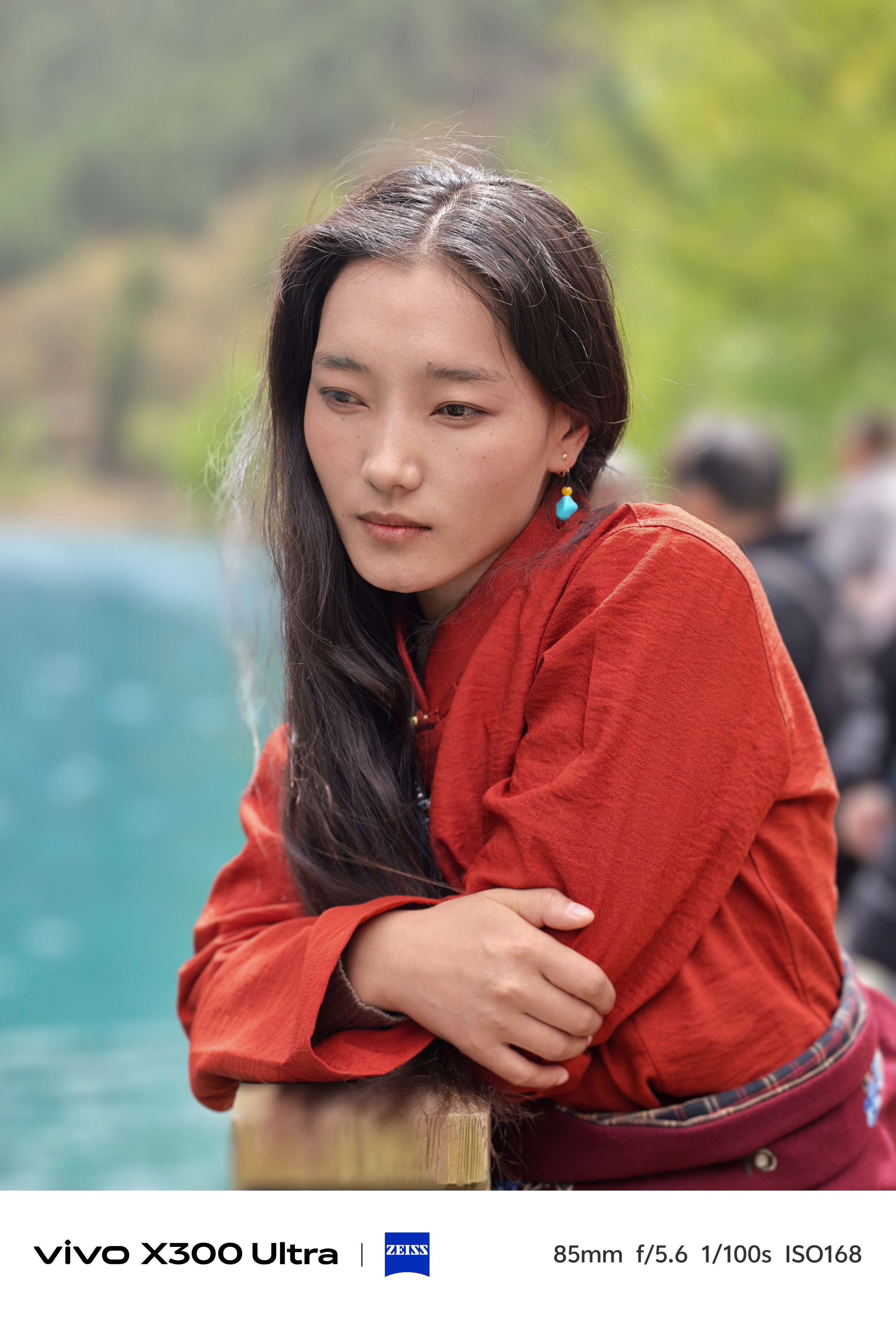 A portrait of a woman with long dark hair wearing a burnt-orange tunic, looking thoughtfully away next to blue water.