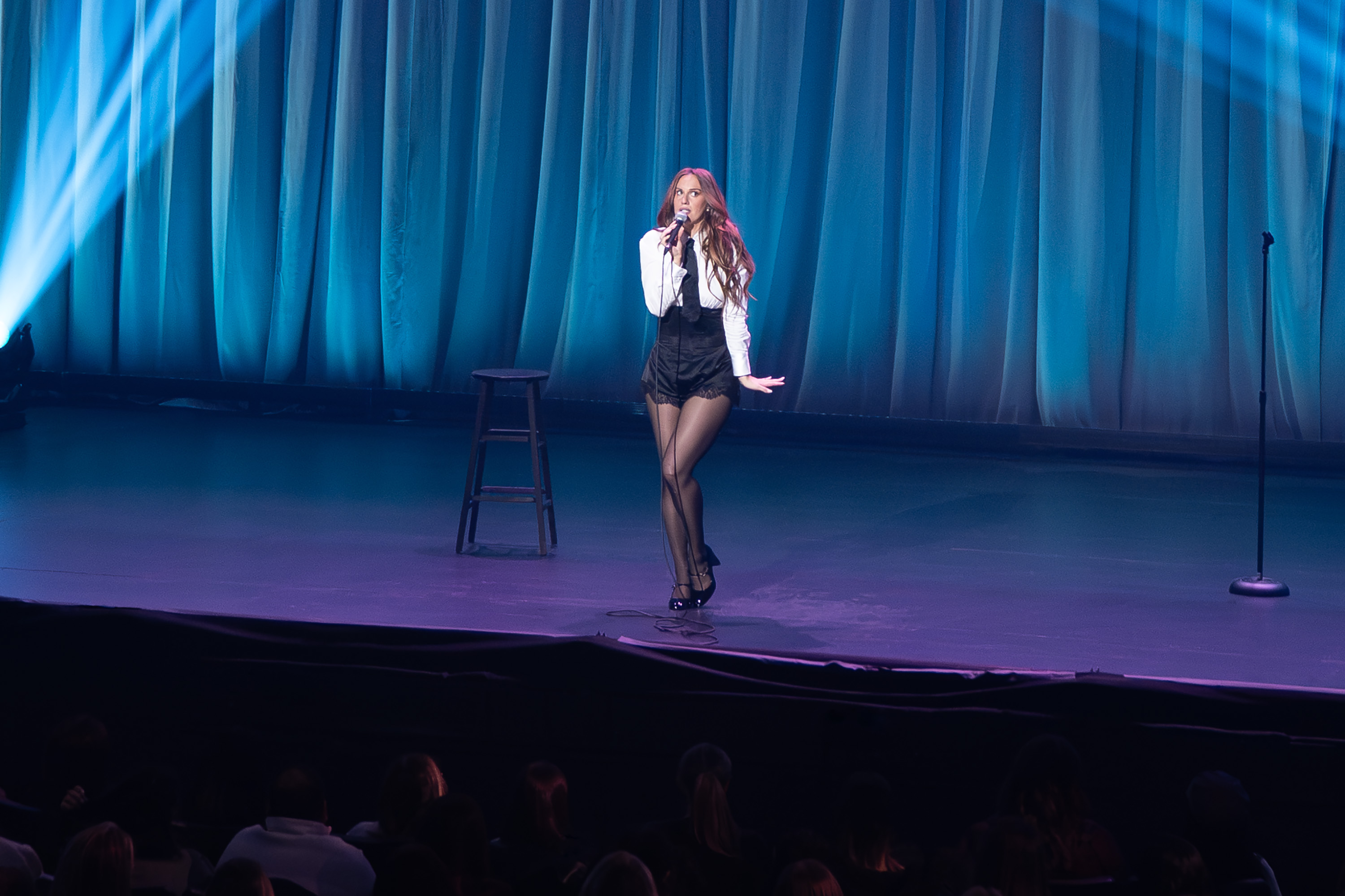 hannah berner wears a white shirt tie black skirt and tights as she poses on stage of her none of my business comedy special standing in front of a blue curtain
