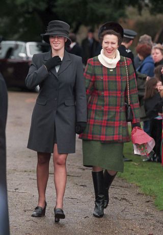 Princess Anne wearing a red and green plaid coat and green skirt with black boots walking next to Zara Tindall in a gray coat and hat with crowds of fans lining the road