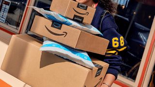 A smiling young woman in a football jersey holding a tower on five Amazon packages and envelopes