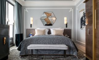 Hotel bedroom with grey bedding and carpet, wooden wardrobe and sideboard and hexagonal mirror over the bed