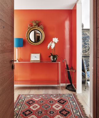 Hallway with a wall painted a bright orange with a matching rug in orange, metallic console table and umbrella stand, round mirror positioned above console table