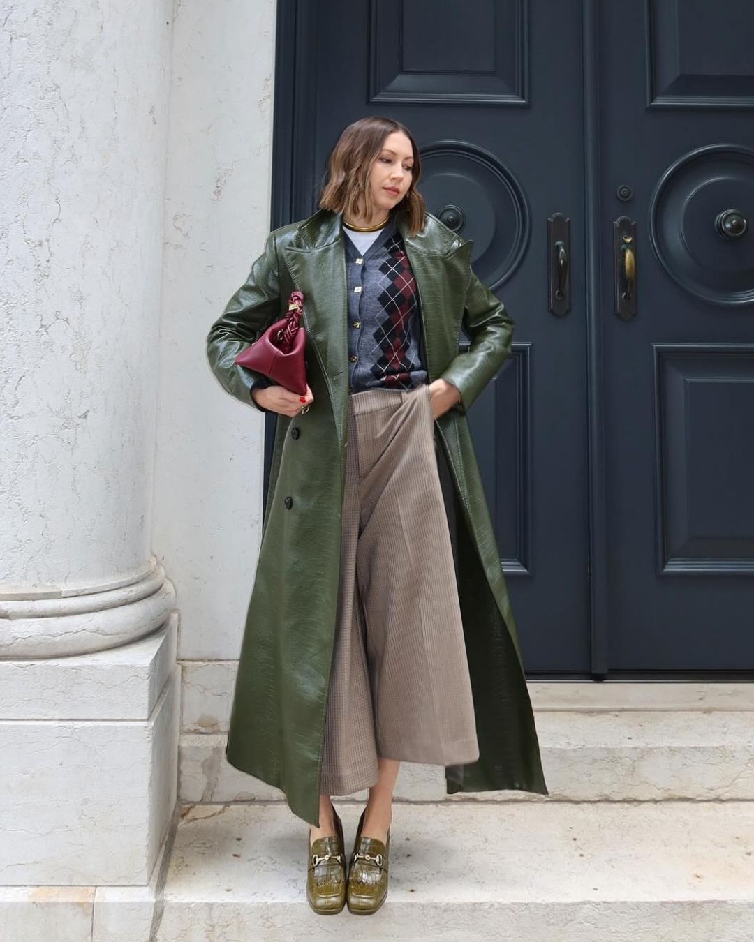 Influencer @trendingwithtinsley stands outside on a step wearing long beige culottes with snakeskin shoes, and longline leather khaki trench coat and an argyle cardigan.