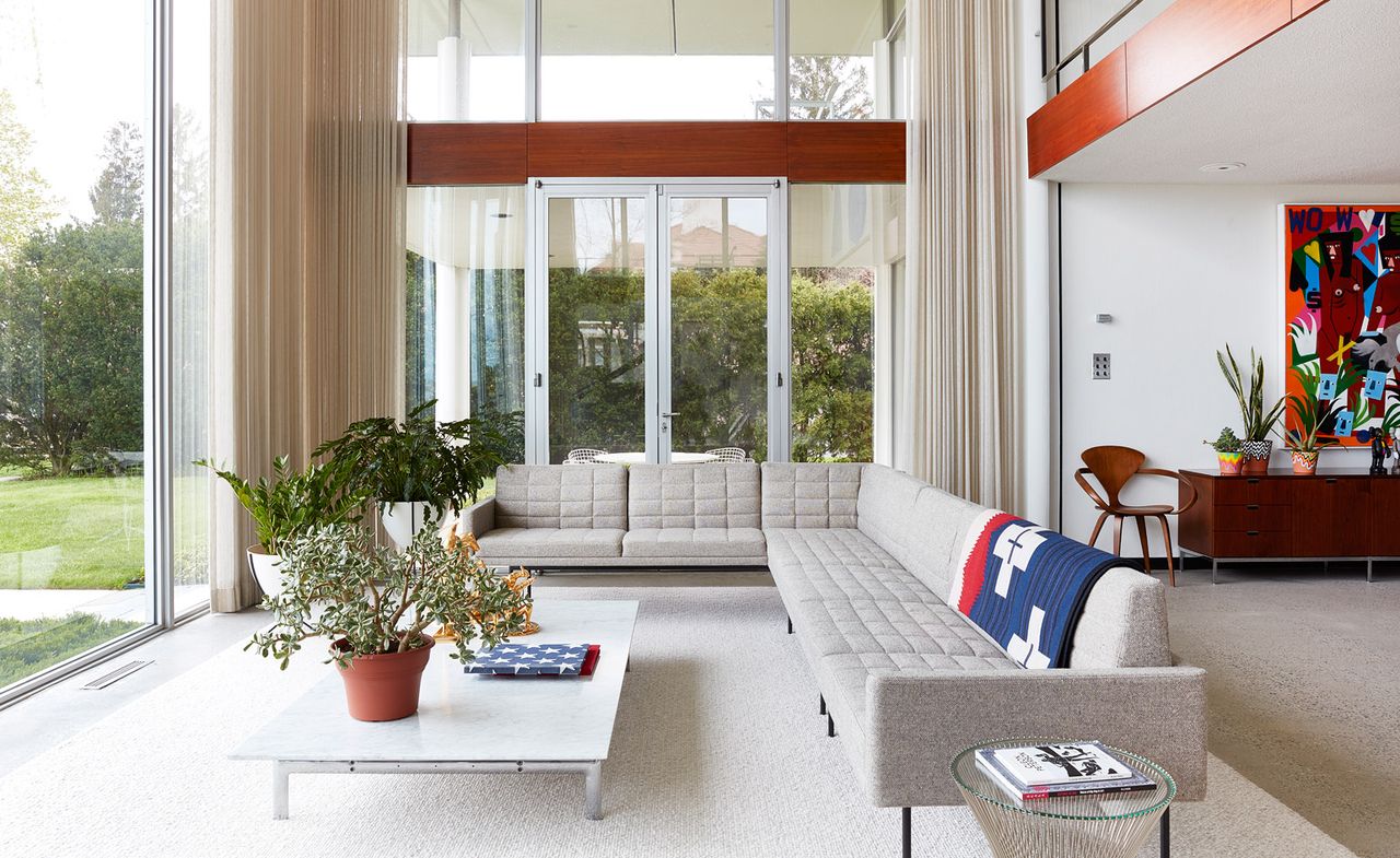 A living room with large windows, coffee table and a grey L-shaped sofa with blue, red and white blanket