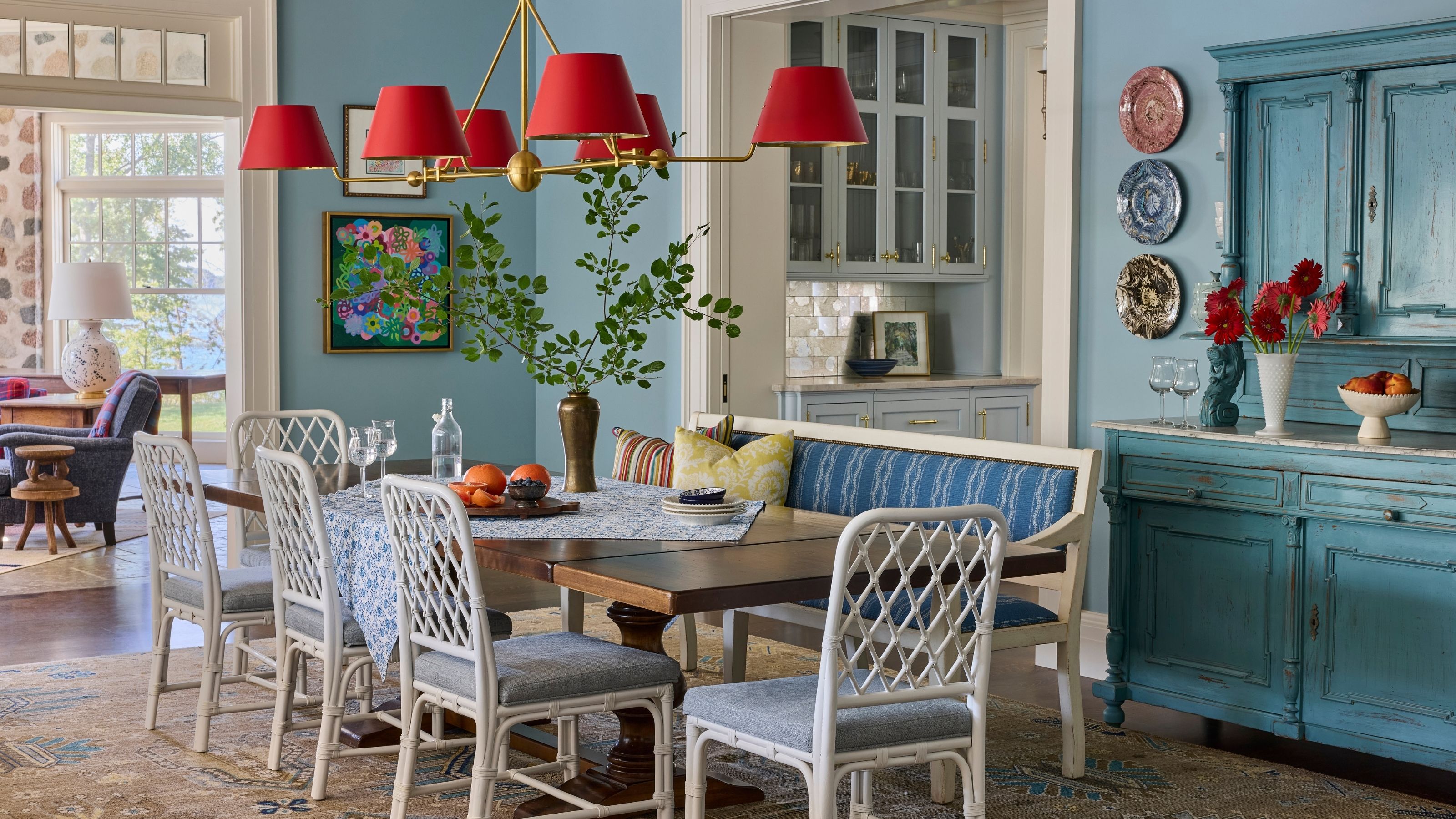 Light blue dining room featuring a long wooden table with white lattice chairs and a blue banquette, illuminated by a striking brass chandelier with vibrant red lampshades.