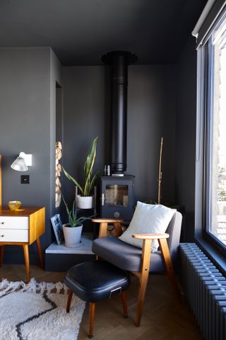 Jason Traves house: A dark and moody seating area in a loft conversion with an armchair and footstool, woodburning stove and deep grey painted walls