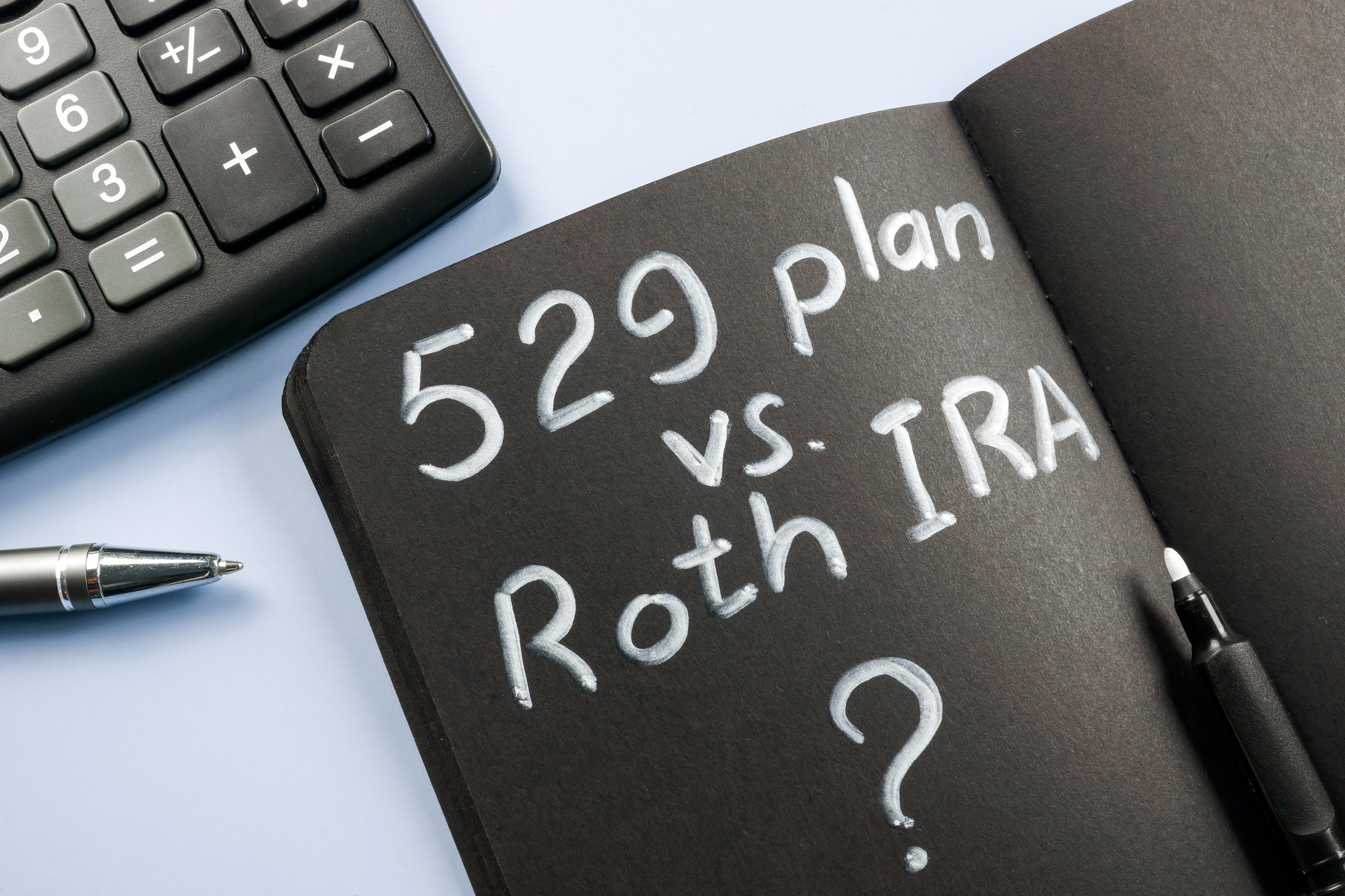 black notebook that says &quot;529 plan vs. Roth IRA?&quot; with pen and calculator nearby