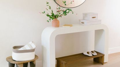 An organized entryway with a wood side table and a woven basket with a striped towel beside a curved white table with a vase of flowers, a pair of storage boxes, and a book below a round wall mirror. Underneath is a wooden bench with a pair of indoor slippers.