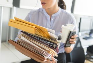 Workplace stress: A woman carrying coffee, a phone and lots of folders in an office