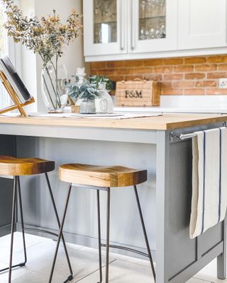 A small kitchen island idea with a grey butcher's block with legs, wood countertops, wood bar stools with metal legs and exposed brick walls