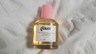 A close up picture of a bottle of the GISOU Honey Infused Hair Perfume Floral Edition - Wild Rose laid on white, textured bedding