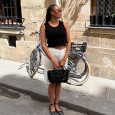 A woman wearing a pair of white jean shorts, a knit black top, and a black bag. 