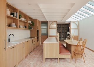 a timber kitchen with terracotta floors. In the middle of the kitchen is a countertop and bench/seating area which leads onto the dining table. There are also four chairs by the table. The kitchen also has open shelving with plates, plants and books stacked inside.