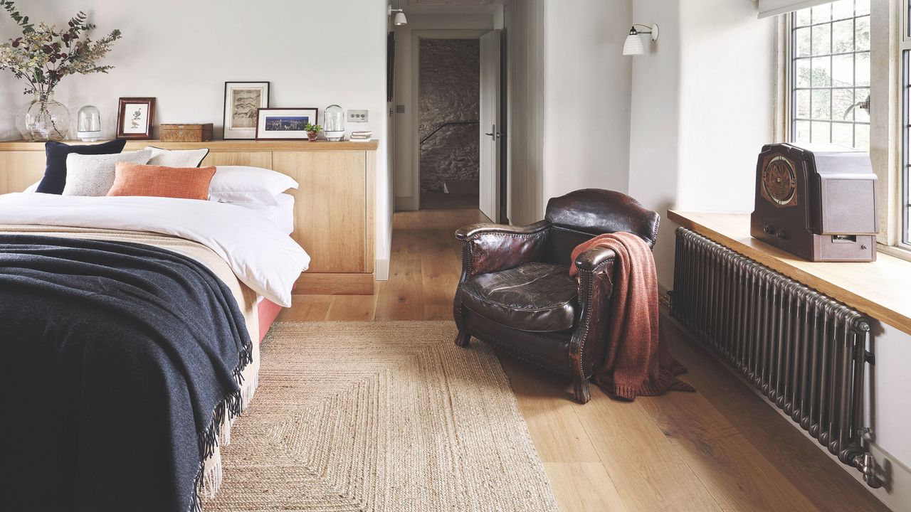 Bedroom with bed on the left, a jute rug on the wooden floor, and a leather armchair by the window next to a vintage radiator