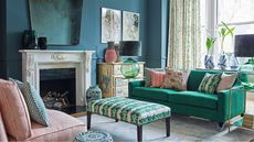 Living room with green sofa and assorted cushions, a traditional decorative fireplace and hearth, padded ottoman, ceramic seat, painted furniture piece and teal walls.