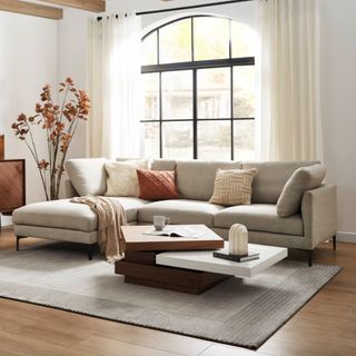 Castlery living room decor styled with floor to ceiling voile curtain panels on a black framed arched window and L-shape sofa with abstract layered wooden coffee table dried tall orange plant in floor vase