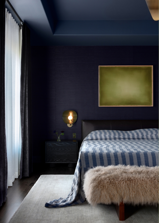 A blue bedroom with a side table, bedside wall-mounted lighting and framed artwork above the bed