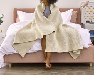 A cream cotton-ribbed weighted blanket wrapped around the shoulders of a woman sat on an upholstered bed