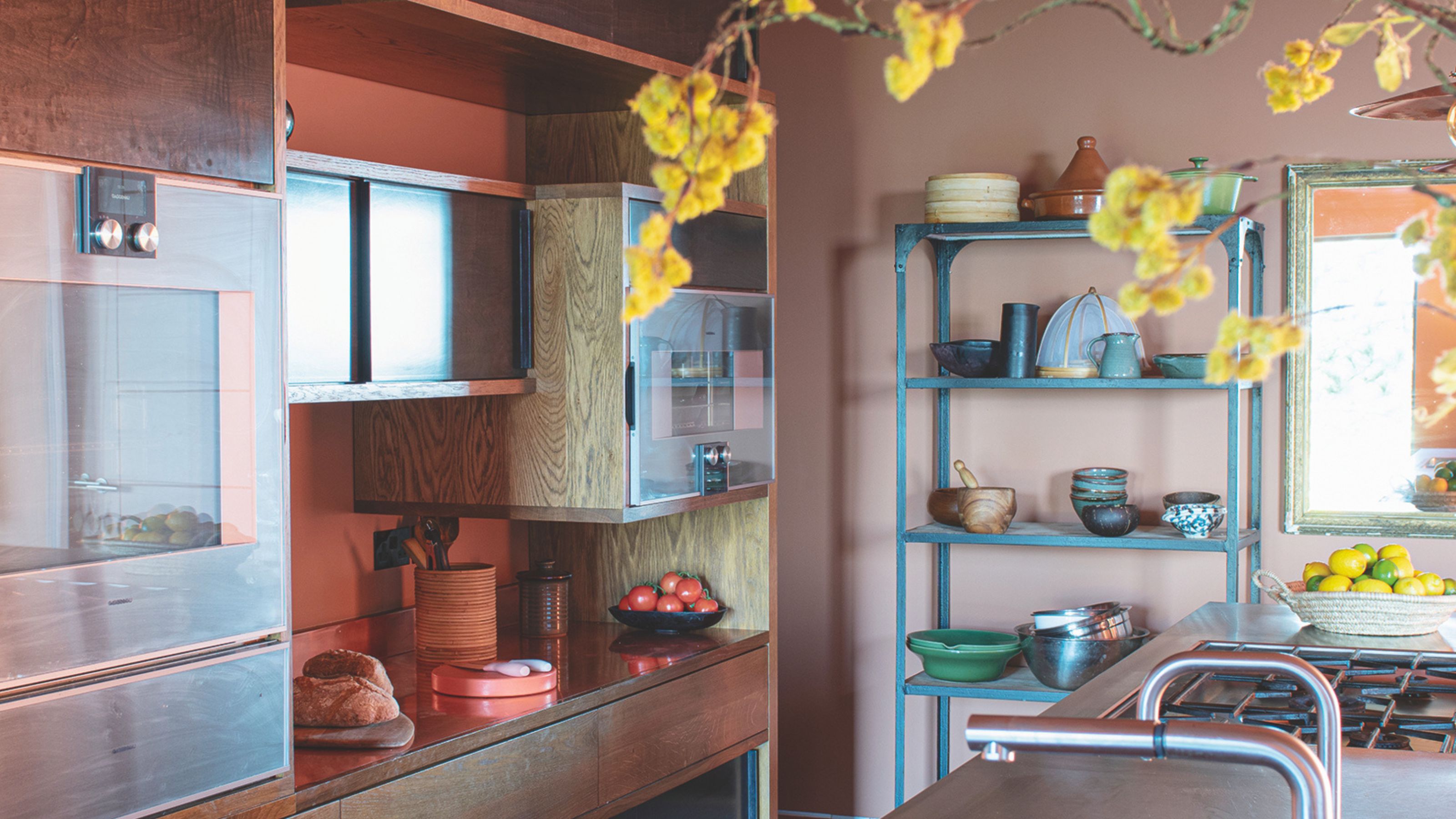 A Japanese style kitchen with bronze accents and yellow blossom just out of view on the kitchen island near the ceiling pendant light with blue freestanding shelves with crockery