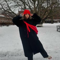 Influencer @tia_dewitt wears a black coat with a red beanie and red knit over her shoulder in the snow.