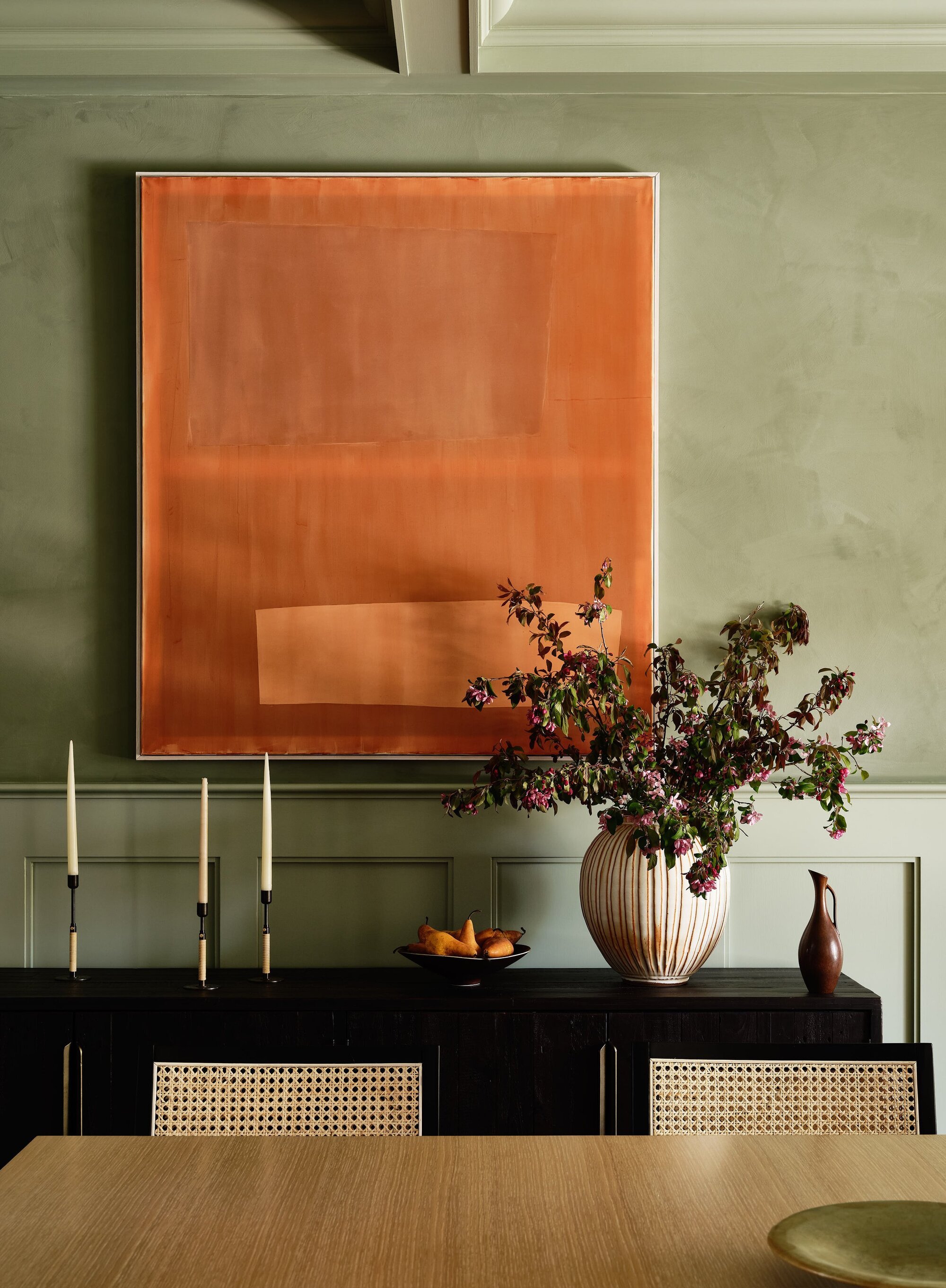 Image of a dining room that is painted a sage green. There is a large, orange painting on the wall with a black console table underneath. On the table is a vase with purple flowers and three candle stick holders with candles in them.
