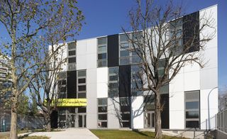 Daytime, outside view of the Lilas Animation Centre in Paris, white building with multiple windows, bare trees, yellow signage to front door entrance, grass lawn, white pathway, clear blue sky