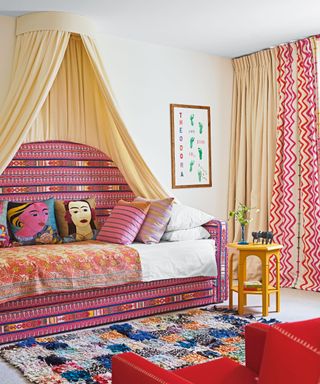Colorful bedroom with unique sofa bed with hanging canopy above, cream carpet, patterned rug, red armchair, cream, red and pink curtains