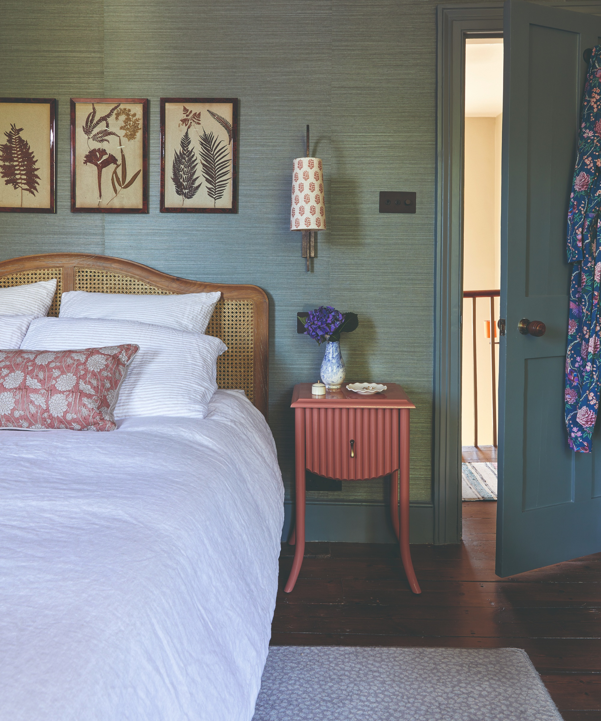 Green textured wallpaper in bedroom with floral artwork on walls, cane bedhead and red bedside table and white bedding