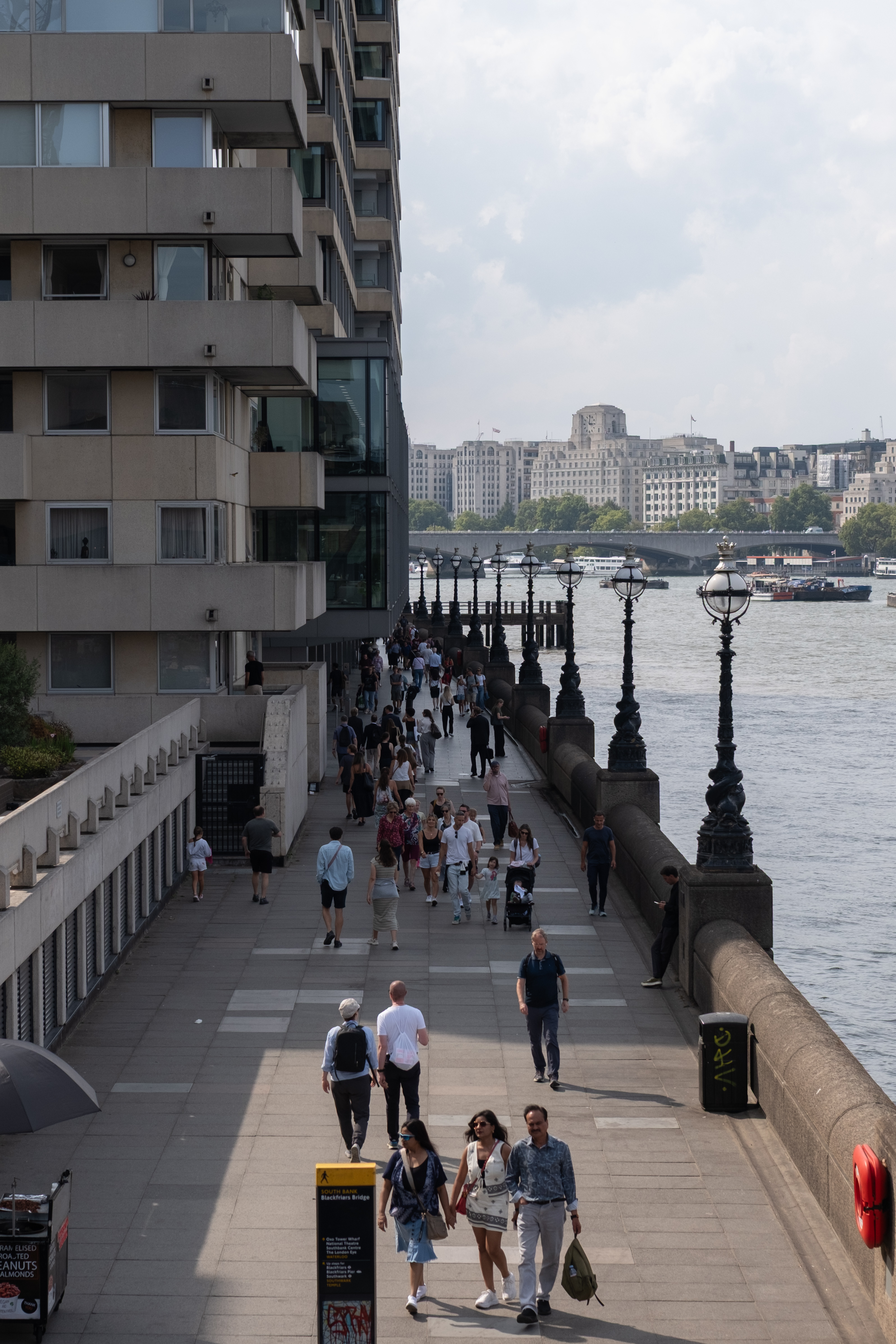 Photograph of a busy South Bank walkway in London beside the River Thames, with pedestrians, ornate lamp posts, and buildings across the river in view.