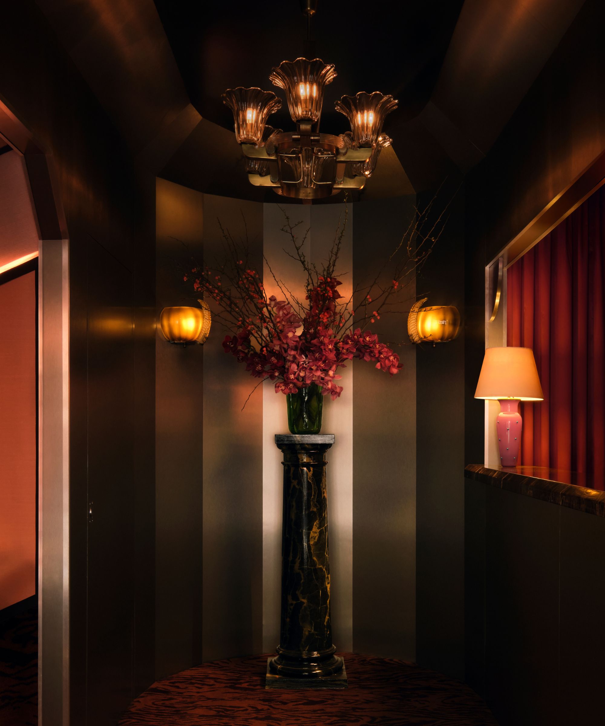 A dramatic, dimly lit hallway featuring a marble pedestal with a large arrangement of pink flowers and branches. The space is illuminated by a vintage glass chandelier, glowing wall sconces, and a pink table lamp against dark, metallic-paneled walls