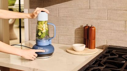 A blue and clear plastic tumbler blender in a neutral kitchen, being operated by a Caucasian woman. 