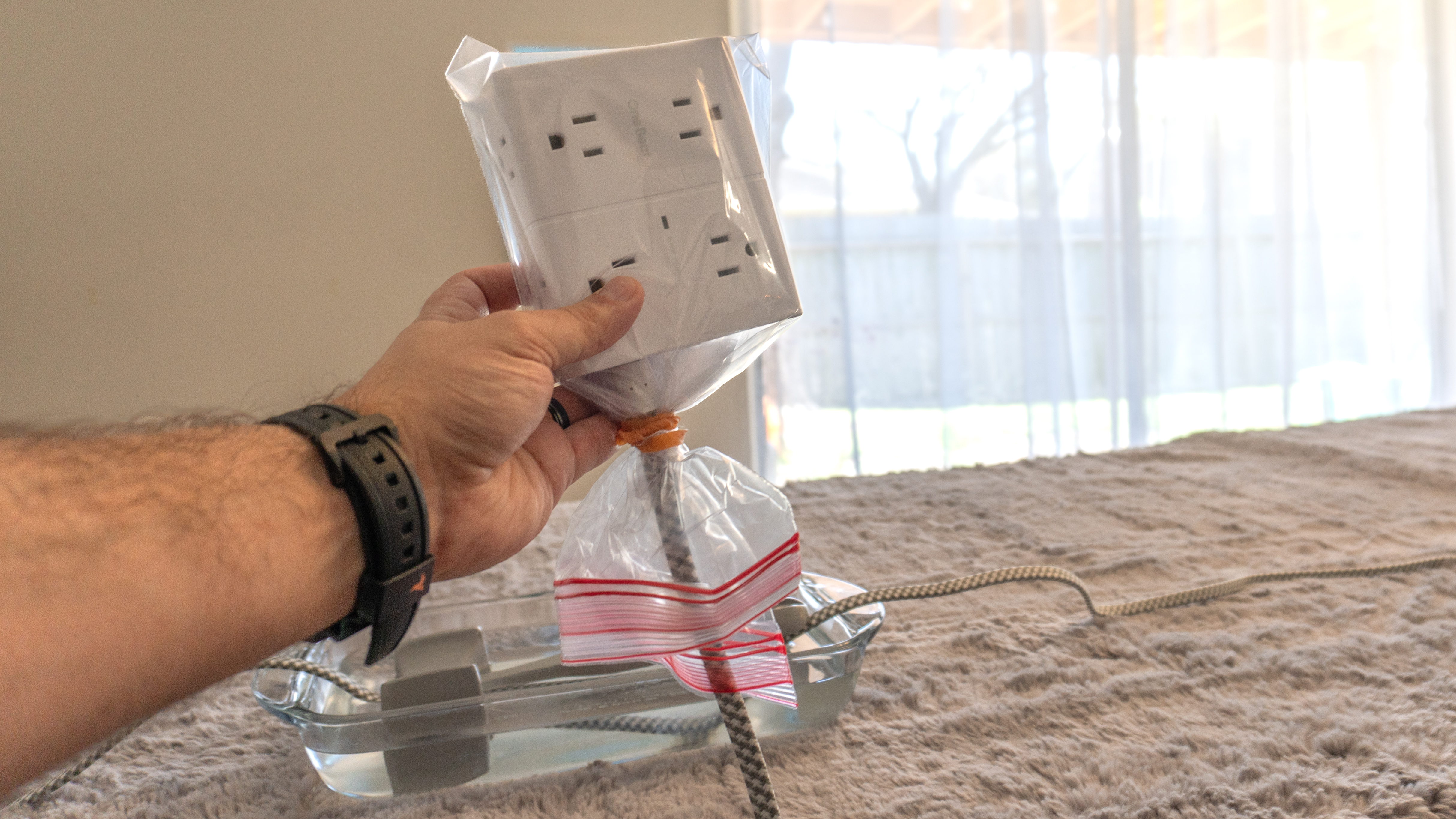 A person holding a surge protector wrapped in a plastic bag in hand while its braided cable is submerged in water in the background