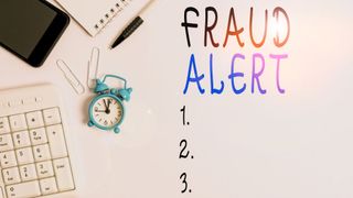 Words 'fraud alert' written on a whiteboard alongside numbers, an alarm clock and a pen.