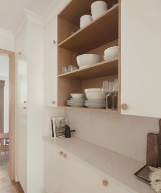 A white narrow kitchen with an open cabinet with wooden circular handles, with white dinnerware and a white countertop with books, trays, and a chopping board on it