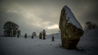 a number of stones stand up in the snow. visible at far left is a tree in the distance. At far right is one of the stones up close with snow on the top of it