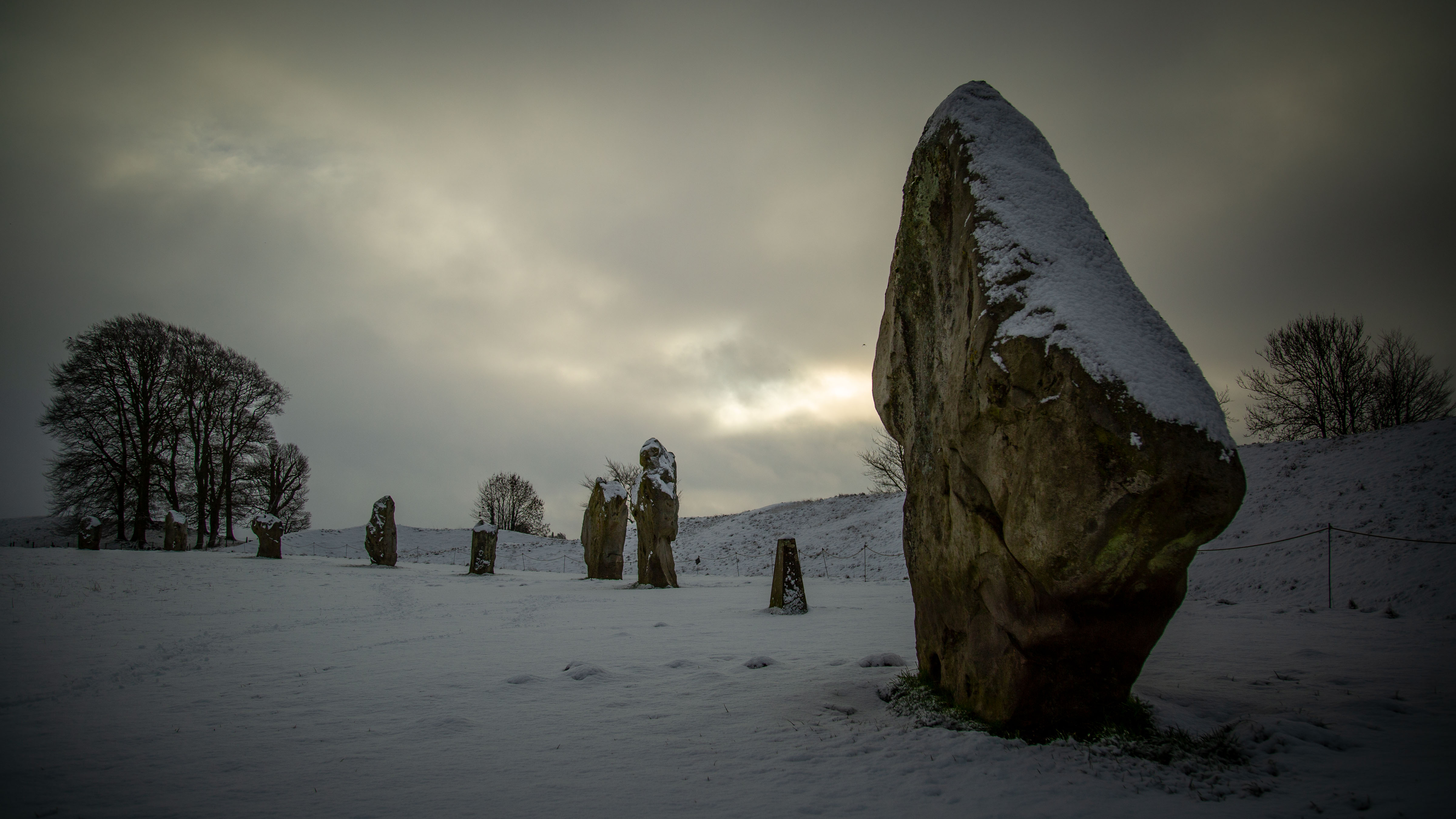 a number of stones stand up in the snow. visible at far left is a tree in the distance. At far right is one of the stones up close with snow on the top of it