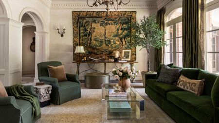 neutral living room with traditional architecture, a vintage tapestry hung on the wall with a dark green velvet sofa, green linen armchairs, an olive tree and large windows flanked by olive green curtains